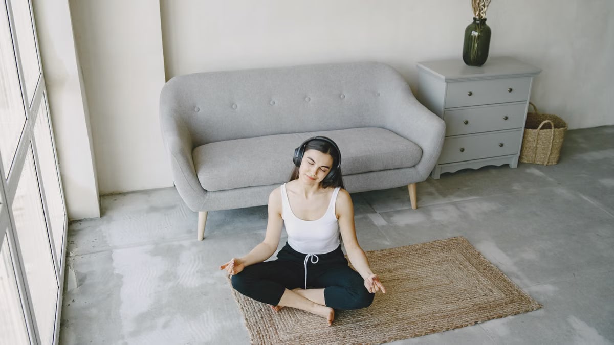 A young woman sits on a mat in a contemporary living room, meditating with headphones on.